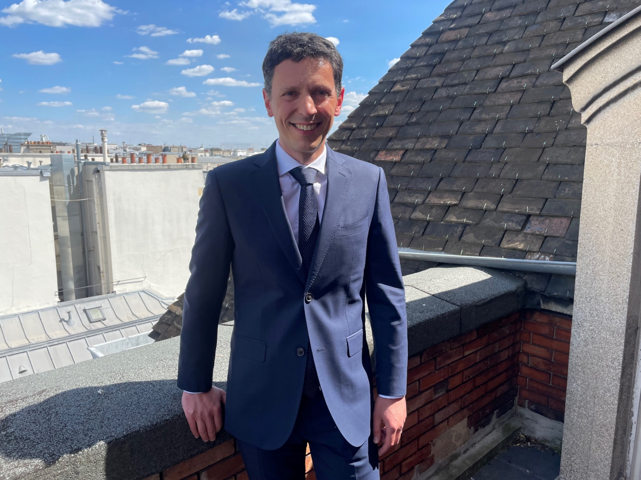 Ein Mann im Anzug steht auf der Terrasse oben auf einem Gebäude mit blauem Himmel im Hintergrund.