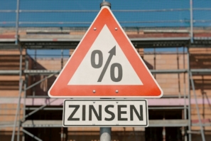 Verkehrschild mit Prozentzeichen, das nach oben weist und dem Wort Zinsen darunter, vor einem Haud im Rohbau.
