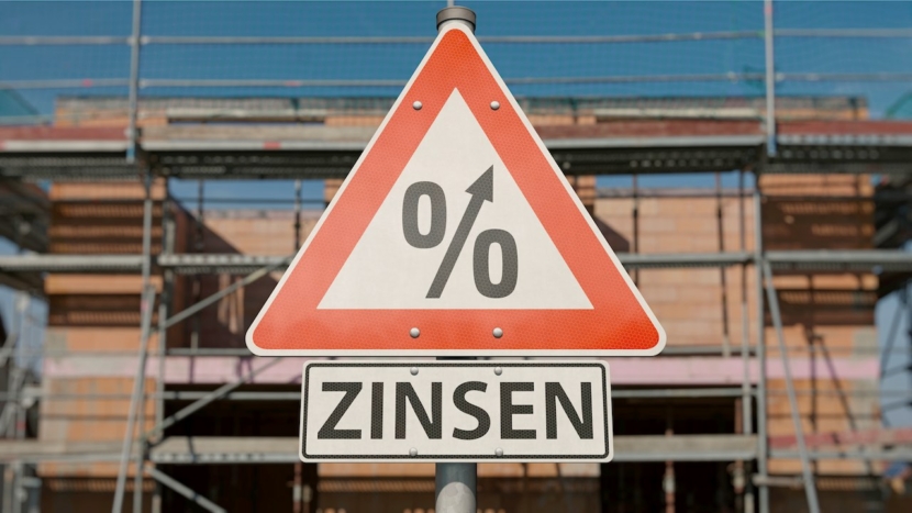 Verkehrschild mit Prozentzeichen, das nach oben weist und dem Wort Zinsen darunter, vor einem Haud im Rohbau.