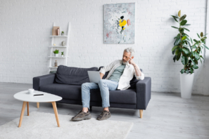 Mann mit grauen Haaren benutzt Laptop, während er auf Couch neben Tasse Kaffee und Smartphone mit leerem Bildschirm auf Couchtisch sitzt